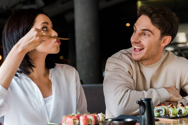 Messa a fuoco selettiva della donna che tiene le bacchette vicino al viso mentre siede con l'uomo allegro nel sushi bar — Foto stock