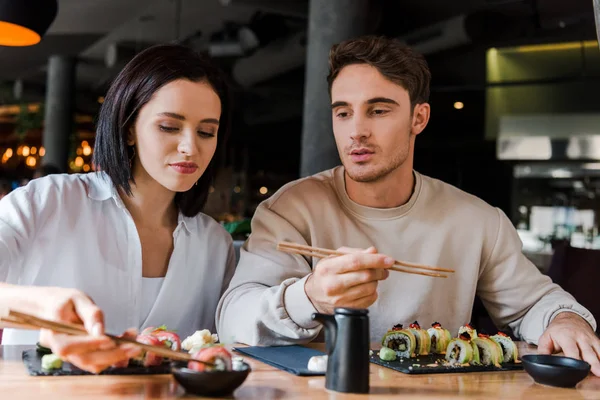 Избирательный фокус привлекательной женщины, держащей палочки с вкусными суши рядом с мужчиной в ресторане — стоковое фото