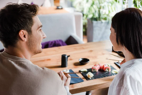 Enfoque selectivo del hombre sosteniendo palillos cerca de la mujer joven en el bar de sushi - foto de stock