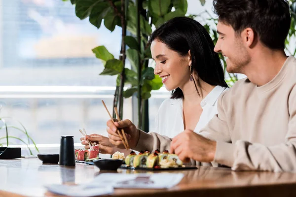 Foco seletivo do jovem segurando pauzinhos e olhando para a mulher feliz no bar de sushi — Fotografia de Stock