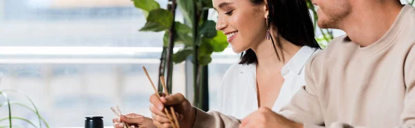 Панорамный снимок молодого человека, держащего палочки для еды рядом с счастливой женщиной в суши-баре — стоковое фото