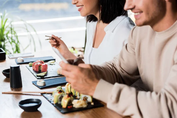 Обрезанный вид молодой женщины, держащей палочки для еды рядом с суши и мужчина в суши-баре — стоковое фото
