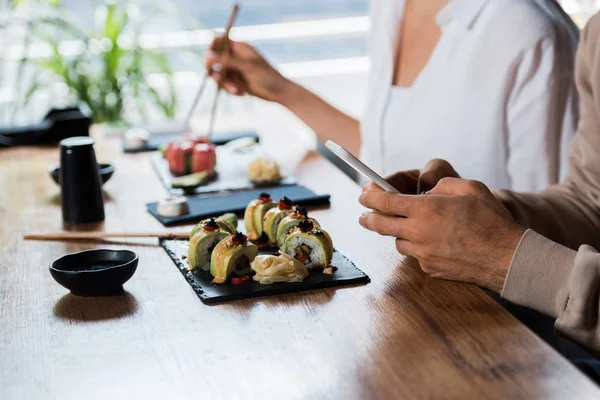 Ausgeschnittene Ansicht eines Mannes mit Smartphone in der Nähe einer jungen Frau mit Essstäbchen in einer Sushi-Bar — Stockfoto