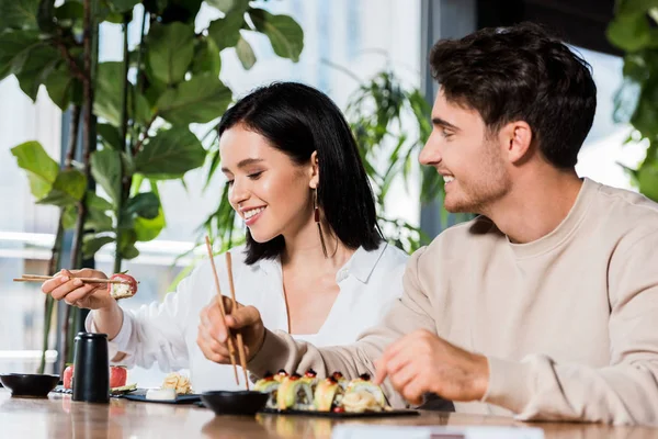 Selective focus of happy man looking at girl holding chopsticks with sushi in restaurant — Stock Photo
