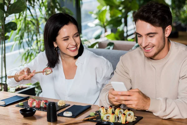 Glückliche Frau hält Essstäbchen mit Sushi in der Hand und schaut fröhlichen Mann mit Smartphone an — Stockfoto