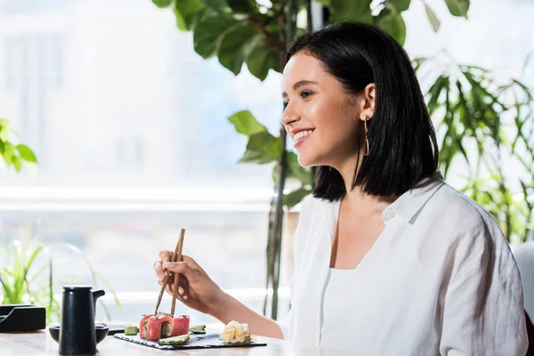 Jovem mulher feliz segurando pauzinhos perto de delicioso sushi no restaurante — Fotografia de Stock