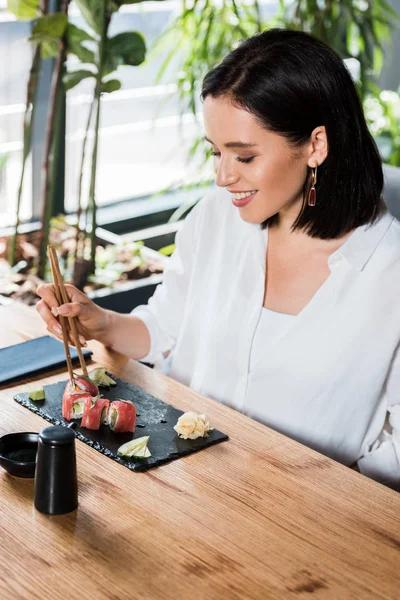 Молодая счастливая женщина держит палочки для еды рядом с вкусными суши в ресторане — стоковое фото