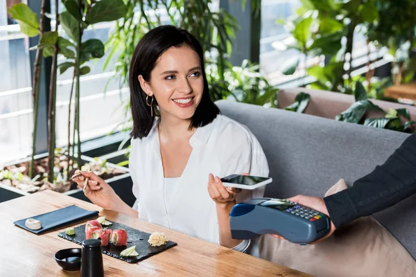 Vista recortada del camarero sosteniendo lector de tarjetas de crédito cerca de mujer alegre pagando con teléfono inteligente - foto de stock