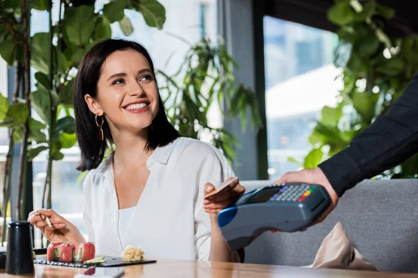 Ausgeschnittene Ansicht des Kellners mit Kreditkartenlesegerät neben lächelnder Frau, die mit Smartphone bezahlt — Stockfoto