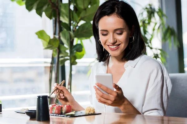 Donna felice utilizzando smartphone e tenendo le bacchette vicino al sushi — Foto stock