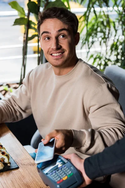 Ausgeschnittene Ansicht einer Kellnerin mit Zahlungsterminal in der Nähe eines glücklichen Mannes mit Smartphone — Stockfoto