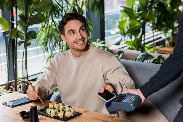 Vista cortada de garçonete segurando terminal de pagamento perto de homem alegre com smartphone — Fotografia de Stock