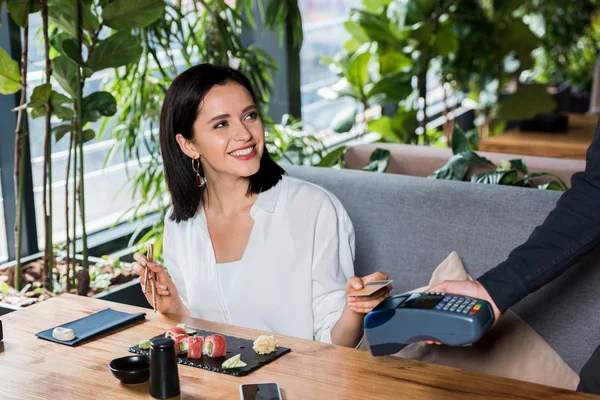 Обрезанный вид официанта, держащего читателя кредитных карт рядом с привлекательной женщиной, платящей кредитной картой — стоковое фото