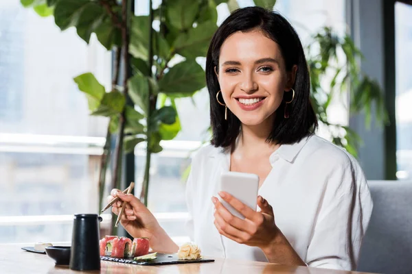 Mujer feliz usando teléfono inteligente cerca de sushi sabroso en el restaurante - foto de stock