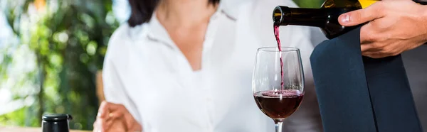 Tiro panorâmico de garçom derramando vinho tinto em vidro perto da mulher — Fotografia de Stock