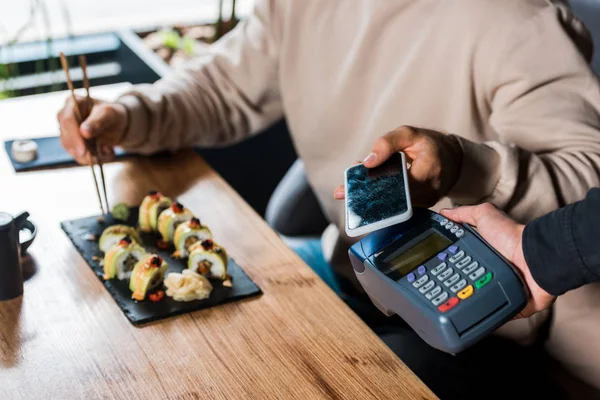 Vista recortada de camarera sosteniendo terminal de pago cerca de hombre con teléfono inteligente en sushi bar - foto de stock