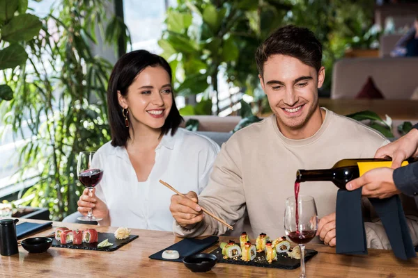 Обрізаний вид на офіціанта, що висипає червоне вино в склянку біля чоловіка і жінки в суші-барі — стокове фото