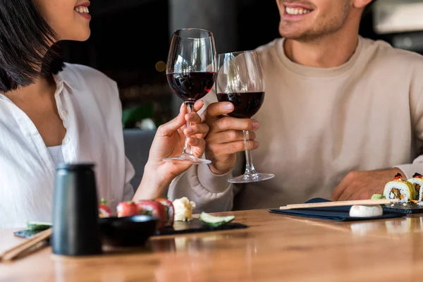 Vista recortada de hombre feliz y mujer alegre tintineo vasos con vino tinto cerca de sushi - foto de stock