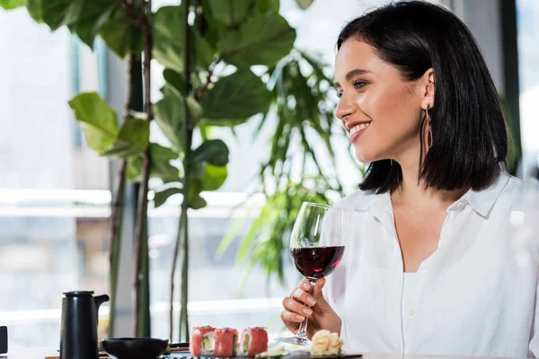 Happy woman holding glass with red wine near tasty meal in sushi bar — Stock Photo