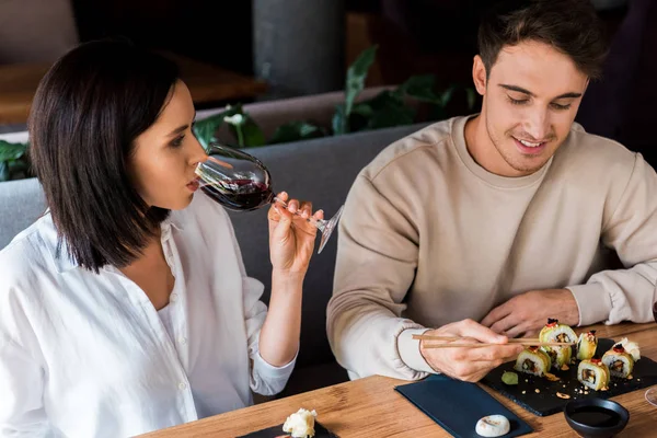 Hombre feliz sosteniendo palillos cerca de sushi mientras la mujer bebe vino - foto de stock