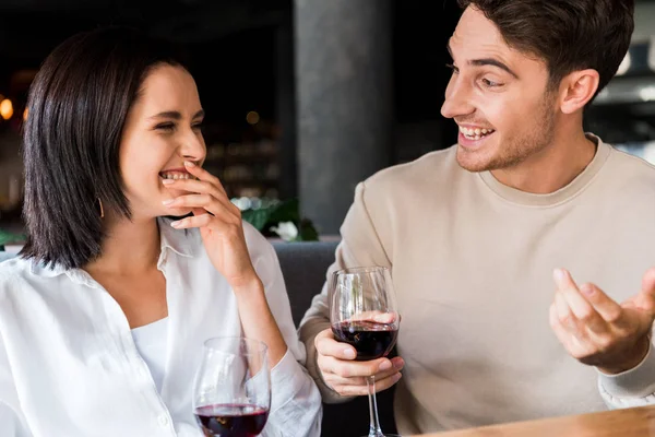 Hombre feliz mirando a la mujer riendo mientras sostiene vasos con vino tinto - foto de stock