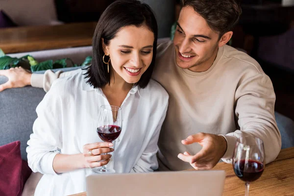 Счастливый мужчина показывает пальцем на ноутбук рядом с женщиной с бокалом вина — стоковое фото