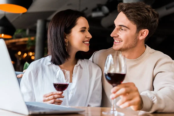 Feliz hombre y mujer con copa de vino mirándose el uno al otro cerca del portátil - foto de stock