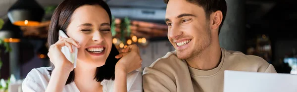 Tiro panorámico de hombre feliz cerca de mujer alegre hablando en el teléfono inteligente - foto de stock