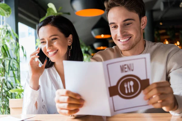 Messa a fuoco selettiva di uomo felice che tiene il menu vicino alla donna che parla sullo smartphone — Foto stock