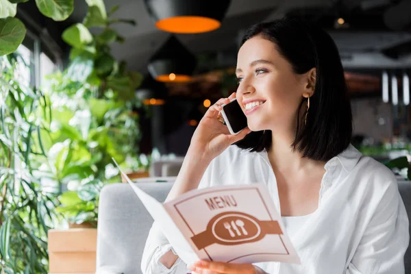 Mujer feliz hablando en el teléfono inteligente y menú de celebración en el restaurante - foto de stock