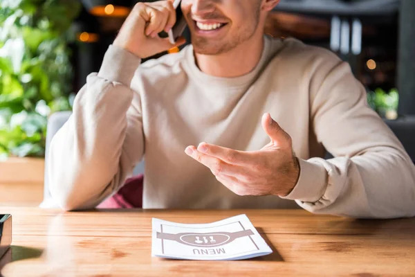 Vista recortada de hombre feliz gesto mientras habla en el teléfono inteligente cerca del menú en la mesa — Stock Photo