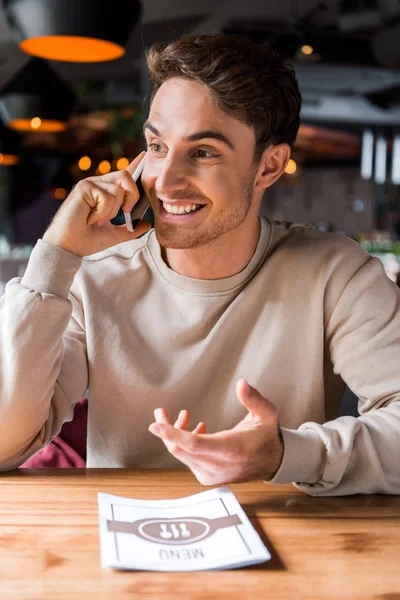 Homme heureux geste tout en parlant sur smartphone près du menu sur la table — Photo de stock