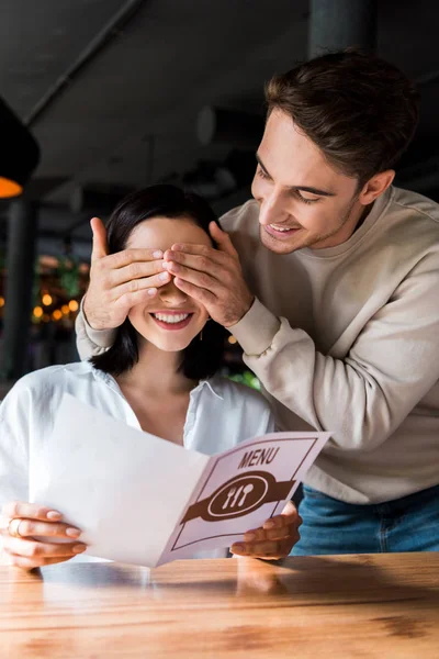 Glücklicher Mann schließt Augen einer fröhlichen Frau, die im Restaurant ein Menü hält — Stockfoto