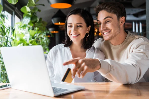 Enfoque selectivo de la mujer feliz mirando hombre portátil con tarjeta de crédito y señalando con el dedo - foto de stock