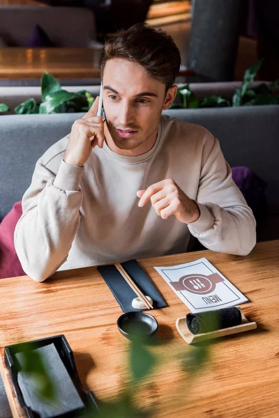 Foco seletivo de homem bonito falando no smartphone no bar de sushi perto da mesa com menu — Fotografia de Stock