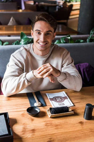 Homem feliz sentado no bar de sushi perto da mesa com menu — Fotografia de Stock