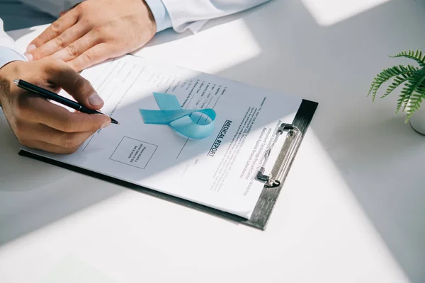 Vista recortada de la escritura del médico en el informe médico cerca de cinta de conciencia azul - foto de stock