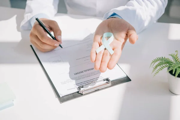 Cropped view of doctor holding blue awareness ribbon while writing in medical report — Stock Photo