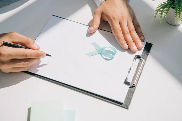 Cropped view of doctor writing diagnosis on clipboard near blue awareness ribbon — Stock Photo