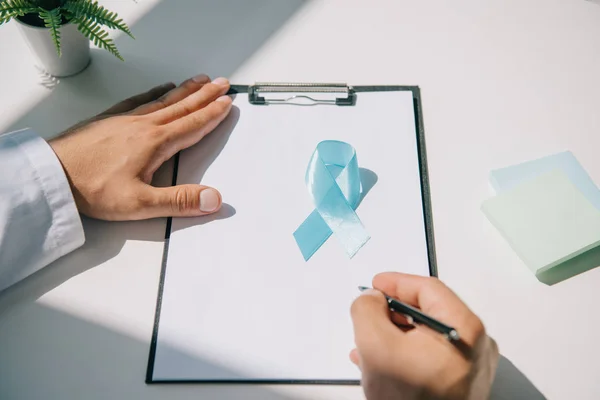 Partial view of doctor writing diagnosis on clipboard near blue awareness ribbon — Stock Photo