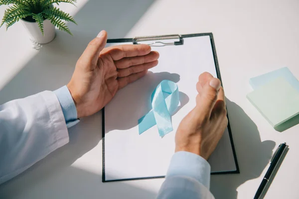 Vista parcial del médico cogido de la mano cerca de la cinta de conciencia azul en el portapapeles con papel en blanco - foto de stock