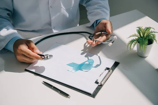 Partial view of doctor holding stethoscope near blue awareness ribbon on clipboard with blank paper — Stock Photo