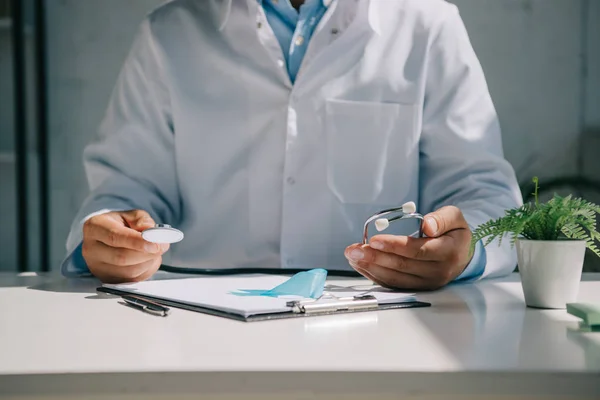 Vista ritagliata del medico seduto alla scrivania e tenendo lo stetoscopio vicino al nastro blu di consapevolezza — Foto stock