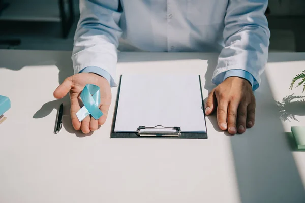 Partial view of doctor holding blue awareness ribbon near clipboard with blank paper — Stock Photo