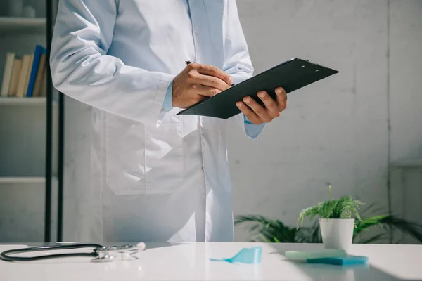 Partial view of doctor writing diagnosis on clipboard while standing near desk — Stock Photo