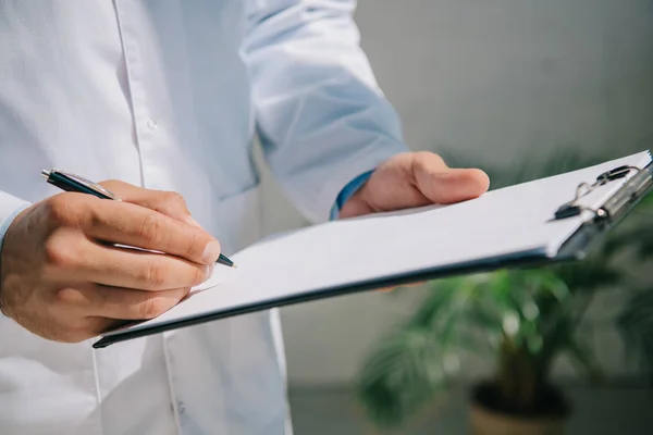 Обрезанный взгляд врача в белом халате, пишущего диагноз в буфер обмена — стоковое фото