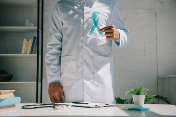 Visão parcial do médico em casaco branco segurando fita de consciência azul enquanto está perto da mesa — Fotografia de Stock