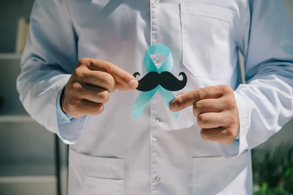 Partial view of doctor holding blue awareness ribbon and paper cut mustache — Stock Photo