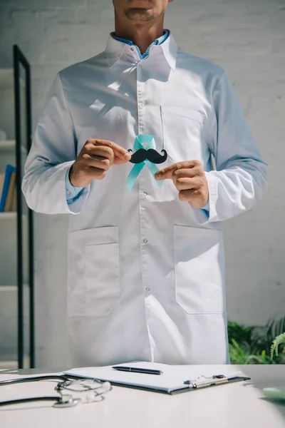 Partial view of doctor in white coat holding blue awareness ribbon with paper cut mustache — Stock Photo