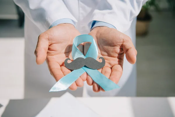 Partial view of doctor holding blue awareness ribbon with paper cut mustache — Stock Photo